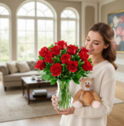 Mujer joven sostiene un jarrón de cristal con un ramo de rosas rojas y un oso de peluche marrón claro en una sala de estar luminosa con sofá y grandes ventanales al fondo