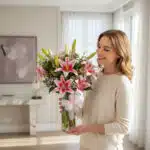 Mujer sonriente con suéter beige sostiene un jarrón de cristal con lirios rosas y flores blancas en un salón luminoso