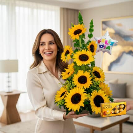 Mujer joven sonriente sostiene un arreglo floral en forma de torre con girasoles amarillos y un globo de estrella con el texto Feliz Día, mientras una mano le entrega una caja de chocolates Ferrero Rocher en una sala de estar moderna y luminosa.