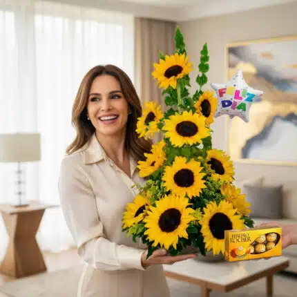 Mujer joven sonriente sostiene un arreglo floral en forma de torre con girasoles amarillos y un globo de estrella con el texto Feliz Día, mientras una mano le entrega una caja de chocolates Ferrero Rocher en una sala de estar moderna y luminosa.