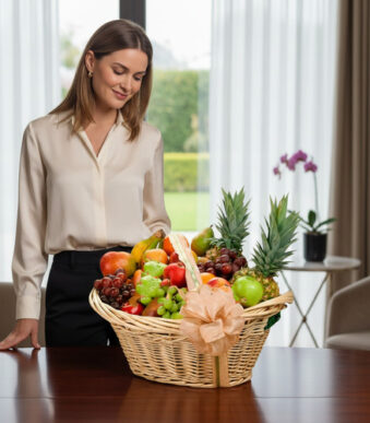 Mujer joven de pie frente a una canasta de mimbre llena de frutas variadas sobre una mesa en un salón luminoso