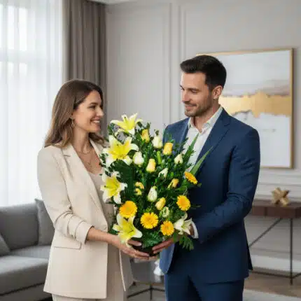 Hombre y mujer vestidos de forma elegante sostienen juntos un gran arreglo de flores amarillas y blancas en una sala de estar moderna y luminosa
