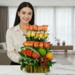 Mujer joven sonriendo detrás de un arreglo floral vertical con rosas naranjas y amarillas combinado con manzana verde, manzana roja, naranja y uvas sobre una base de tallos verdes en una cocina moderna