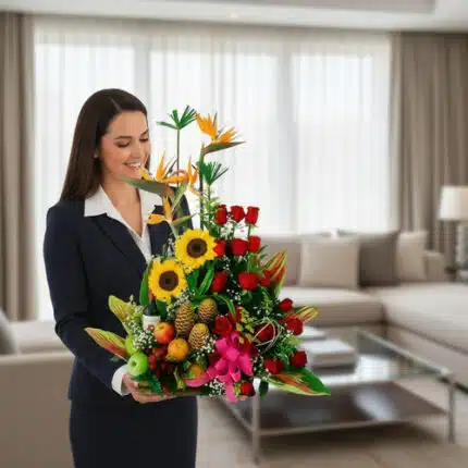 Mujer vestida de traje oscuro sosteniendo un gran arreglo de flores tropicales con girasoles, rosas rojas y frutas como piñas y manzanas en una sala de estar luminosa