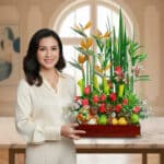 Mujer con blusa crema sostiene un centro de mesa de madera con flores tropicales de colores y frutas variadas en un interior luminoso