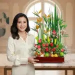 Mujer con blusa crema sostiene un centro de mesa de madera con flores tropicales de colores y frutas variadas en un interior luminoso