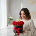 Mujer joven sonriendo mientras sostiene una caja redonda negra llena de rosas rojas con lazo rojo, en interior luminoso