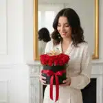 Mujer joven de cabello castaño ondulado con blusa crema sosteniendo una caja redonda negra con rosas rojas y lazo rojo, de pie en un salón blanco con espejo dorado y chimenea clásica al fondo