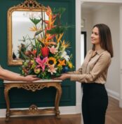 Mujer joven de pie en una sala elegante recibe un gran arreglo floral tropical con lirios, girasoles, aves del paraíso y frutas sobre una bandeja