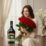 Joven sonriente con vestido claro sostiene un gran ramo de rosas rojas junto a una botella de licor oscuro sobre una mesa en un salón elegante