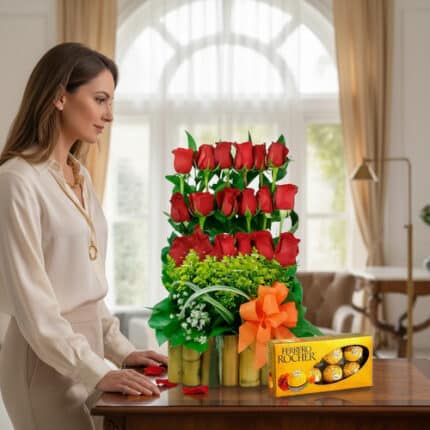 Mujer observando un arreglo de rosas rojas en varios niveles con follaje verde en base de bambú y caja de chocolates Ferrero Rocher sobre una mesa de madera en sala iluminada