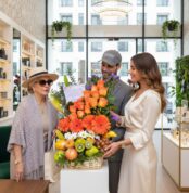 Tres personas observan una canasta grande con flores naranjas y frutas variadas en el interior de una tienda moderna y luminosa