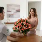 Mujer sonriendo recibe de un repartidor un gran arreglo de rosas y lirios color salmón en una canasta sobre una mesa de madera en una sala moderna