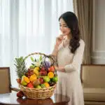 Mujer joven de pie junto a una canasta de mimbre llena de frutas frescas como piña, uvas, naranjas, kiwis y manzanas sobre una mesa en una sala elegante