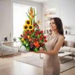 Mujer joven con vestido beige sosteniendo un arreglo de flores y frutas coloridas en una sala de estar moderna y luminosa