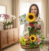 Mujer sonriente sosteniendo una canasta de regalo con girasoles, rosas rojas, lirios, frutas y un globo que dice feliz cumpleaños en una sala elegante