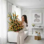 Mujer joven de pie en una sala de estar moderna sosteniendo un gran arreglo de flores naranjas y verdes en una base rectangular oscura