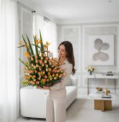 Mujer joven de pie en una sala de estar moderna sosteniendo un gran arreglo de flores naranjas y verdes en una base rectangular oscura