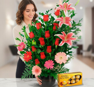 Mujer sonriendo junto a un arreglo floral grande con rosas rojas, lirios rosados y gerberas en maceta negra sobre mesa de mármol, acompañada de una caja de chocolates Ferrero Rocher