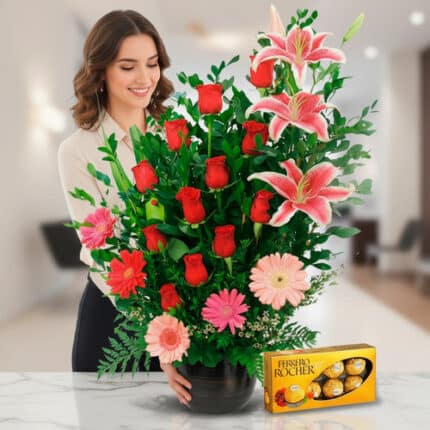Mujer sonriendo junto a un arreglo floral grande con rosas rojas, lirios rosados y gerberas en maceta negra sobre mesa de mármol, acompañada de una caja de chocolates Ferrero Rocher