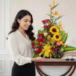 Mujer joven sonriente recibe un arreglo floral con girasoles, rosas rojas, aves del paraíso y frutas tropicales sobre una base verde en interior elegante