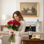 Mujer joven con vestido claro abrazando un ramo de rosas rojas junto a una mesa con botella de vino tinto y caja de bombones en una sala de estar elegante