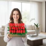 Mujer joven sonriente sostiene un arreglo rectangular de rosas rojas con follaje verde en una sala de estar luminosa y moderna