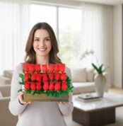 Mujer joven sonriente sostiene un arreglo rectangular de rosas rojas con follaje verde en una sala de estar luminosa y moderna