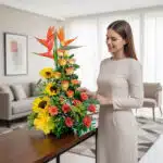 Mujer joven con vestido beige acomodando un arreglo floral alto con girasoles, rosas naranjas y aves del paraíso sobre una mesa en una sala moderna