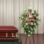 Arreglo floral fúnebre con lirios rosas y rosas blancas sobre caballete al lado de un ataúd de madera barnizada en una sala con cortinas claras