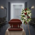 Ataúd de madera barnizada con detalles metálicos plateados junto a corona funeraria de flores blancas y rojas en una sala de velatorio desenfocada