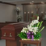 Arreglo de flores blancas con lazo morado frente a un ataúd de madera en una capilla funeraria
