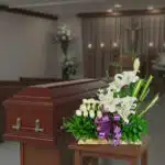 Arreglo de flores blancas con lazo morado frente a un ataúd de madera en una capilla funeraria