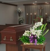 Arreglo de flores blancas con lazo morado frente a un ataúd de madera en una capilla funeraria