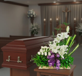 Arreglo de flores blancas con lazo morado frente a un ataúd de madera en una capilla funeraria