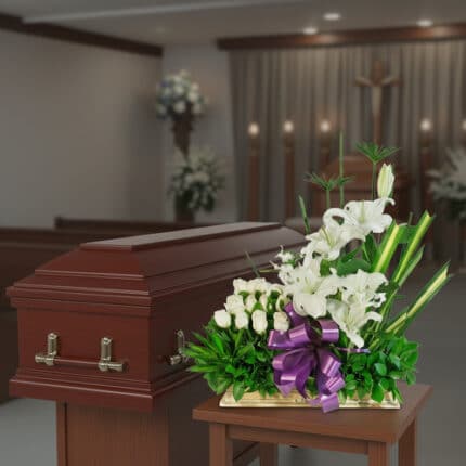 Arreglo de flores blancas con lazo morado frente a un ataúd de madera en una capilla funeraria