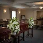 Sala de velatorio interior con dos ataúdes de madera oscura decorados con arreglos de flores blancas y verdes, rodeados de sillas vacías