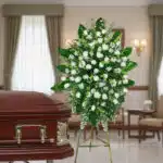 Corona de flores funeraria con rosas blancas y follaje verde sobre caballete metálico al lado de un ataúd de madera barnizada en una sala velatoria
