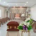 Ataúd de madera barnizada con herrajes dorados junto a arreglo de flores blancas y lazo morado en una sala de velatorio
