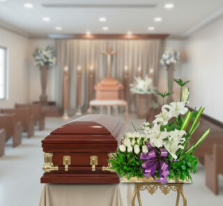 Ataúd de madera barnizada con herrajes dorados junto a arreglo de flores blancas y lazo morado en una sala de velatorio