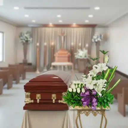 Ataúd de madera barnizada con herrajes dorados junto a arreglo de flores blancas y lazo morado en una sala de velatorio