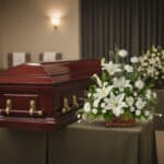 Ataúd de madera color caoba con herrajes dorados junto a un arreglo floral blanco sobre mesa cubierta con mantel en una sala de velatorio