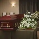Ataúd de madera color caoba con herrajes dorados junto a un arreglo floral blanco sobre mesa cubierta con mantel en una sala de velatorio