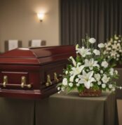 Ataúd de madera color caoba con herrajes dorados junto a un arreglo floral blanco sobre mesa cubierta con mantel en una sala de velatorio