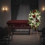 Arreglo floral grande con lirios blancos, rosas rojas y flores blancas sobre caballete de madera, colocado al lado de un ataúd de madera oscura en una sala de velatorio con cortinas y sillas vacías