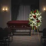 Arreglo floral grande con lirios blancos, rosas rojas y flores blancas sobre caballete de madera, colocado al lado de un ataúd de madera oscura en una sala de velatorio con cortinas y sillas vacías