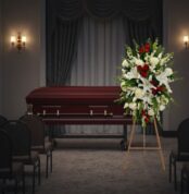 Arreglo floral grande con lirios blancos, rosas rojas y flores blancas sobre caballete de madera, colocado al lado de un ataúd de madera oscura en una sala de velatorio con cortinas y sillas vacías