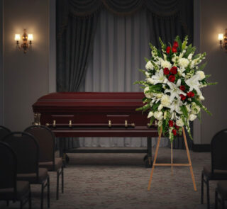 Arreglo floral grande con lirios blancos, rosas rojas y flores blancas sobre caballete de madera, colocado al lado de un ataúd de madera oscura en una sala de velatorio con cortinas y sillas vacías
