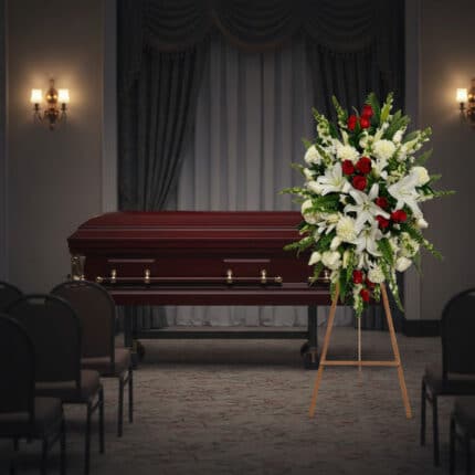 Arreglo floral grande con lirios blancos, rosas rojas y flores blancas sobre caballete de madera, colocado al lado de un ataúd de madera oscura en una sala de velatorio con cortinas y sillas vacías