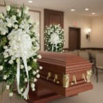 Ataúd de madera pulida con herrajes dorados junto a coronas de flores blancas en una sala de velatorio